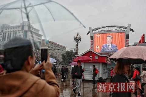 北京站广场上一位市民拿起手机拍摄大屏幕上的北京十九大开幕式直播。